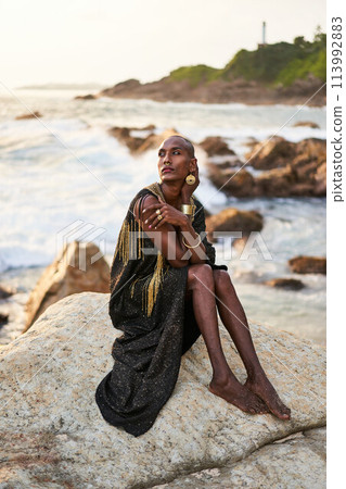 Non-binary black person in luxury dress, golden jewelry on beach rocks in ocean. Trans ethnic fashion model wearing jewellery in posh gown poses gracefully in tropical seaside location on sunset. Non-binary black person in luxury dress, golden jewelry on beach rocks in ocean. Trans ethnic fashion model wearing jewellery in posh gown poses gracefully in tropical seaside location on sunset. 113992883