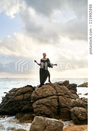Gender fluid black person poses gracefully standing on rocks in ocean . Androgynous ethnic fashion model in posh dress and jewellery on rocky beach by storm. Pride month 113992898