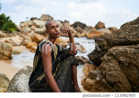 Non-binary ethnic fashion model in luxury dress sits on rocks by ocean. Trans sexual black person in jewellery and posh clothes poses gracefully in tropical seaside location. Pride month. Non-binary ethnic fashion model in luxury dress sits on rocks by ocean. Trans sexual black person in jewellery and posh clothes poses gracefully in tropical seaside location. Pride month. 113992905
