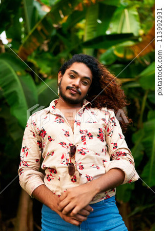 Confident gay man with curly hair stands in tropical setting. Flamboyant pose, bright floral shirt open, denim pants. Arms crossed, smiling, gender expression, lush green foliage backdrop. Confident gay man with curly hair stands in tropical setting. Flamboyant pose, bright floral shirt open, denim pants. Arms crossed, smiling, gender expression, lush green foliage backdrop. 113992931