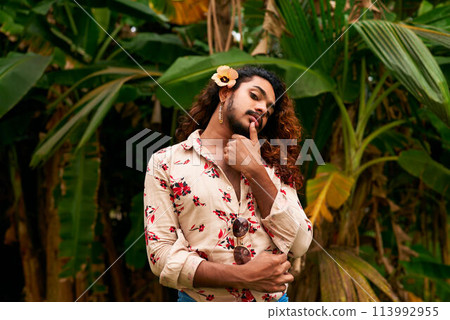 Sri Lankan gay man with a flower in his hair stands in a tropical setting, touching his chin thoughtfully. His floral shirt blends with the rich plants, expressing identity and soft masculinity. 113992955