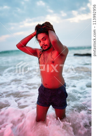 Expressive gay man poses on beach at sunset, waves crash around. Stands confident, hands in hair, expressive gaze, pride, freedom. Wearable art, lifestyle moment. Perfect for LGBT cruises, pride. 113992956