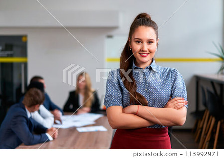 Confident asian exec woman stands in modern office, arms crossed, smiling at camera, team in discussion behind her, embodying female leadership, pro success in a smart casual attire. 113992971