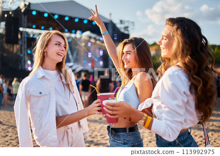 Group of young women cheering with drinks at beach music festival, sunlit stage in background. Smiling females in casual summer attire enjoy live concert with friends on sandy shore. Group of young women cheering with drinks at beach music festival, sunlit stage in background. Smiling females in casual summer attire enjoy live concert with friends on sandy shore. 113992975