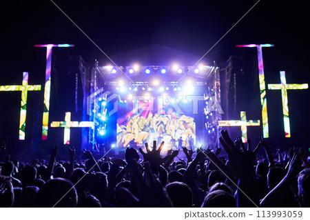 Outdoor religious music festival at night with vibrant stage lights. Excited audience raises hands in worship. Live band performs for believers at Christian event. Unity, praise atmosphere fills air. Outdoor religious music festival at night with vibrant stage lights. Excited audience raises hands in worship. Live band performs for believers at Christian event. Unity, praise atmosphere fills air. 113993059