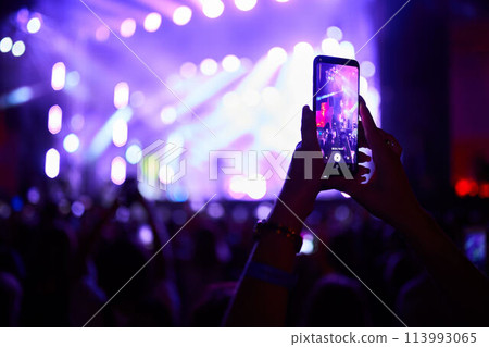 Excited audience at music fest, vibrant lights illuminate stage, fan records live performance on smartphone, friends enjoy night out, band plays in background, festival vibe in summer evening. Excited audience at music fest, vibrant lights illuminate stage, fan records live performance on smartphone, friends enjoy night out, band plays in background, festival vibe in summer evening. 113993065