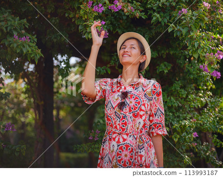 young asian woman stands in sunny green park, admiring blooming bougainvillea bush. The flower's bright colors are beautiful contrast to park's lush greenery. young asian woman stands in sunny green park, admiring blooming bougainvillea bush. The flower's bright colors are beautiful contrast to park's lush greenery. 113993187