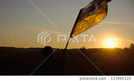 Young man in military uniform waving flag of Ukraine against beautiful sunset at background. Male ukrainian army soldier lifted national banner at countryside. Victory against russian aggression 113995120
