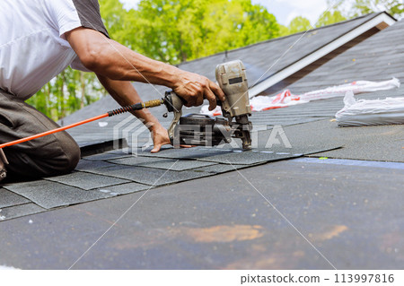 In addition to using an air pneumatic nail gun, roofer installs new asphalt bitumen shingles In addition to using an air pneumatic nail gun, roofer installs new asphalt bitumen shingles 113997816