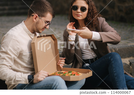 Young bloggers make photo of vegan pizza outdoor. Happy couple take food photography. Man hold pizza box on his knees. Vegan fast food Young bloggers make photo of vegan pizza outdoor. Happy couple take food photography. Man hold pizza box on his knees. Vegan fast food 114000067