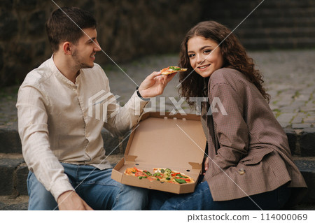 Smiling love couple eating vegan pizza at street on stairs. Handsome man give slice of pizza to woman 114000069