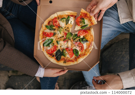 Top view image of couple grab slices of pizza from box at the outdoor. Man and woman hands taking pizza. Vegan pizza with fresh tomatoes basil and broccoli. Lactose and gluten free Top view image of couple grab slices of pizza from box at the outdoor. Man and woman hands taking pizza. Vegan pizza with fresh tomatoes basil and broccoli. Lactose and gluten free 114000072