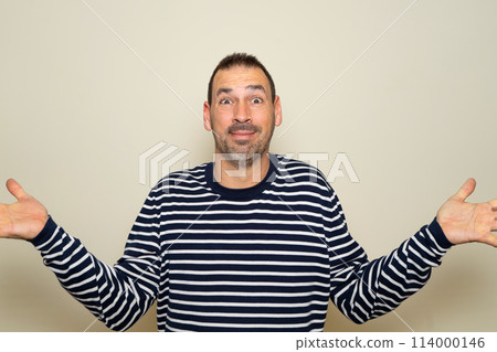 Hispanic man with beard in his 40s wearing a striped sweater clueless and confused with open arms, no idea concept. Isolated on beige studio background. 114000146