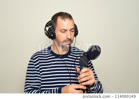 Bearded Hispanic man in his 40s wearing a striped sweater looking confused while inspecting a microphone and headphones, isolated on beige background. 114000149