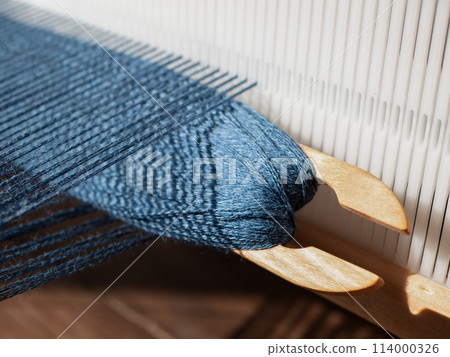 Wooden weaving shuttle with blue yarn is inserted in open shed. Separation between upper and lower warp yarns through which the weft is woven 114000326