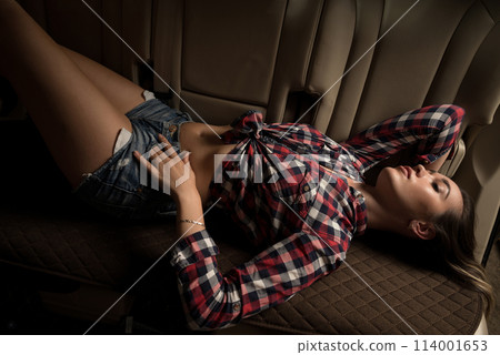 A beautiful girl in a plaid shirt and denim shorts is sitting in the car 114001653