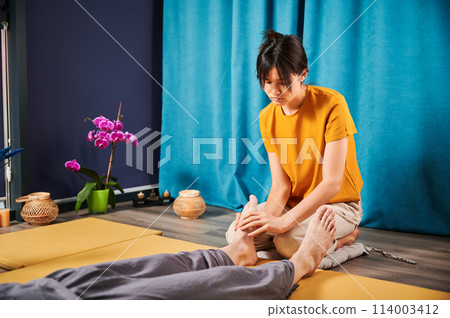 Masseuse making thai yoga massage. Woman therapist doing traditional massage treatment for male patient indoors. Client lying on mat, meditating, enjoying hand massage in calm atmosphere. 114003412
