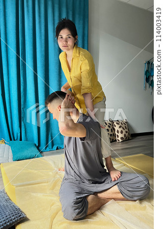 Masseuse making thai yoga massage. Woman therapist doing traditional massage treatment, stretching male patient's body. Client sitting on mat, enjoying hand massage in calm atmosphere. 114003419
