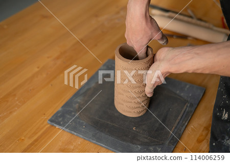 Close-up of a man's hands making a patterned cylinder out of clay. Close-up of a man's hands making a patterned cylinder out of clay. 114006259