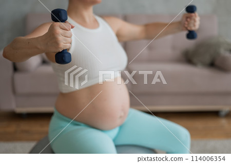 Pregnant woman doing exercises with dumbbells while sitting on a fitness ball at home. Close-up of a pregnant belly. Pregnant woman doing exercises with dumbbells while sitting on a fitness ball at home. Close-up of a pregnant belly. 114006354
