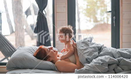 Red-haired Caucasian woman lies in bed with her baby son in a country house with panoramic windows.  114006420