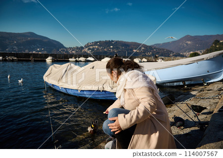 Back view of a young woman in beige coat, sitting nears boats by the Como lake, relaxing in the lakefront and feeding by hands the birds and ducks with bread Back view of a young woman in beige coat, sitting nears boats by the Como lake, relaxing in the lakefront and feeding by hands the birds and ducks with bread 114007567