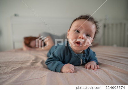 A three-month-old boy lies on his stomach on the bed and his mother sleeps behind him. Postpartum depression. A three-month-old boy lies on his stomach on the bed and his mother sleeps behind him. Postpartum depression. 114008374