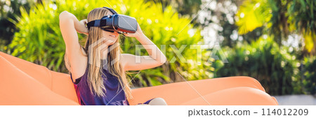 BANNER, LONG FORMAT Summer lifestyle portrait of pretty girl sitting on the orange inflatable sofa and uses virtual reality headset on the beach of tropical island. Relaxing and enjoying life on air BANNER, LONG FORMAT Summer lifestyle portrait of pretty girl sitting on the orange inflatable sofa and uses virtual reality headset on the beach of tropical island. Relaxing and enjoying life on air 114012209