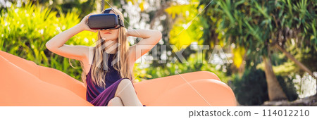 BANNER, LONG FORMAT Summer lifestyle portrait of pretty girl sitting on the orange inflatable sofa and uses virtual reality headset on the beach of tropical island. Relaxing and enjoying life on air 114012210
