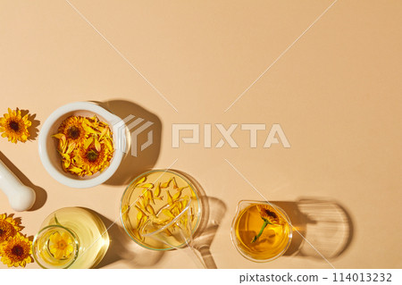 Laboratory glassware filled with liquid and oil extracted from Calendula flowers. A white mortar containing yellow flowers and petals featured. Vacant space for product advertising. Copy space 114013232