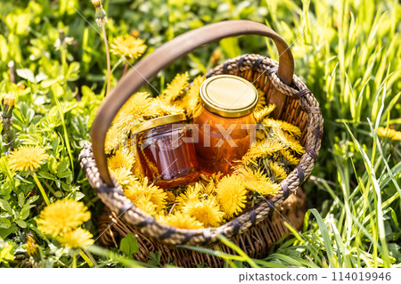 Dandelion flowers in a wicker basket with a jar of honey placed on the ground somewhere in a meadow 114019946