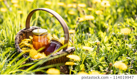 Dandelion flowers in a wicker basket with a jar of honey placed on the ground somewhere in a meadow 114019947