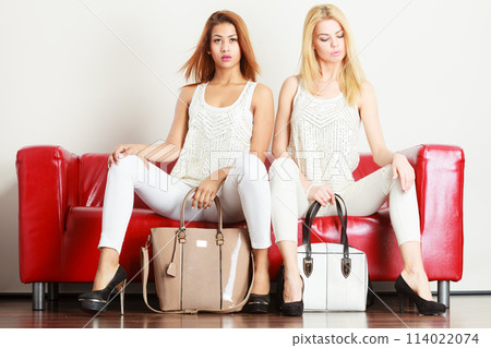 Two women sitting on sofa presenting bags 114022074