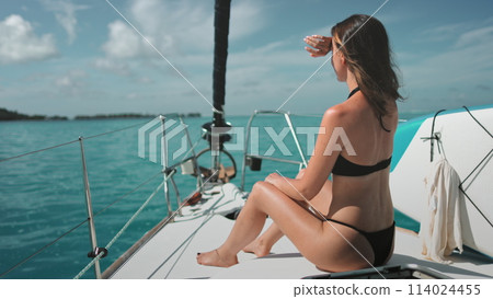 A woman wearing a bikini is seated on a boat, enjoying the view and the sun while on the water. A woman wearing a bikini is seated on a boat, enjoying the view and the sun while on the water. 114024455