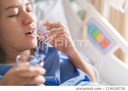 Asian woman patient with medical drip or IV drip drinking water with medicine in hospital ward, Selective focus, Healthcare concept. Asian woman patient with medical drip or IV drip drinking water with medicine in hospital ward, Selective focus, Healthcare concept. 114026738