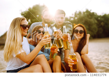 Group of friends cheers and drink beers on the beach. Young friends relaxing and having picnic. Suummertime. 114031774