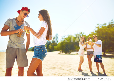Smiling young couple clinking glasses with beer while people barbecuing in the background. 114032849