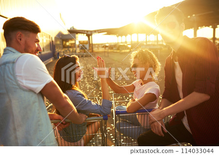 Group of happy young people having fun on shopping trolleys. Multiethnic young people racing on shopping cart. 114033044