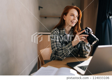 Cheerful female artist adjusting new professional photo camera or choosing photos for editing sitting in front of laptop computer at home office workplace, smiling looking away. Cheerful female artist adjusting new professional photo camera or choosing photos for editing sitting in front of laptop computer at home office workplace, smiling looking away. 114033106