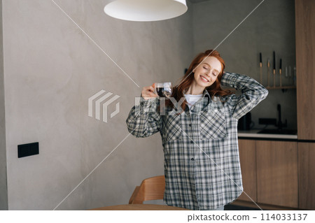 Cheerful young woman enjoying holding cup drinking coffee standing in kitchen on sunny morning. Happy smiling female relaxing on caffeine break alone. Satisfied lady drinking coffee at home. Cheerful young woman enjoying holding cup drinking coffee standing in kitchen on sunny morning. Happy smiling female relaxing on caffeine break alone. Satisfied lady drinking coffee at home. 114033137
