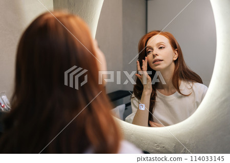 Reflection on mirror of attractive young redhead woman enjoying her appearance while getting ready in bathroom. Happy beautiful female touching face look in bathroom mirror, enjoy healthy skin. Reflection on mirror of attractive young redhead woman enjoying her appearance while getting ready in bathroom. Happy beautiful female touching face look in bathroom mirror, enjoy healthy skin. 114033145