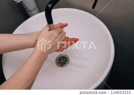 Closeup top view of unrecognizable woman washing dirty hands rubbing using sanitizer soap, rinsing under warm running water above bathroom sink. Concept of hygienic procedures in home bathroom. Closeup top view of unrecognizable woman washing dirty hands rubbing using sanitizer soap, rinsing under warm running water above bathroom sink. Concept of hygienic procedures in home bathroom. 114033153
