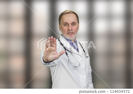 Serious physician doctor shows stop gesture sign. Elder male doctor shows his palm. Blurred checkered windows background. Serious physician doctor shows stop gesture sign. Elder male doctor shows his palm. Blurred checkered windows background. 114034572