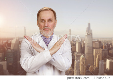 Happy senior medic doctor in white coat shows no reject sign. Urban cityscape background. Happy senior medic doctor in white coat shows no reject sign. Urban cityscape background. 114034606