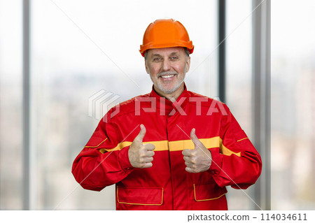 Portrait of a smiling handyman construction worker showing both thumb up. Blurred windows background. Portrait of a smiling handyman construction worker showing both thumb up. Blurred windows background. 114034611