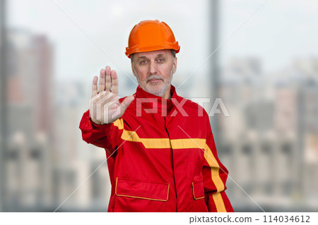 Mature construction worker handyman shows the no reject gesture sign. Blurred indoor windows background. Mature construction worker handyman shows the no reject gesture sign. Blurred indoor windows background. 114034612