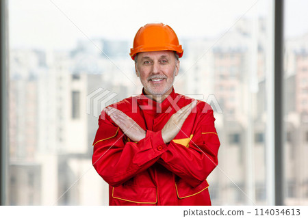 Smiling old caucasian worker shows the sign of prohibition. Bright urbanscape background. Smiling old caucasian worker shows the sign of prohibition. Bright urbanscape background. 114034613