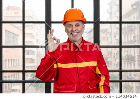 Smiling construction handyman worker in red uniform shows okay gesture sign. Checkered window background. Smiling construction handyman worker in red uniform shows okay gesture sign. Checkered window background. 114034614