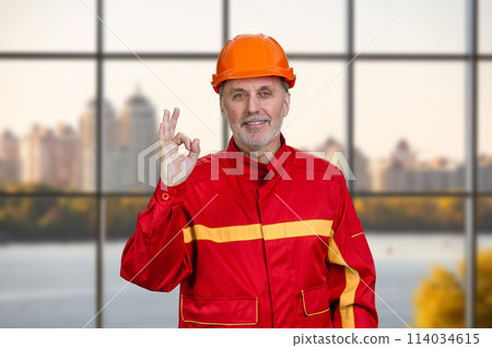 Happy smiling construction worker in red uniform shows okay gesture sign. Checkered window background. 114034615