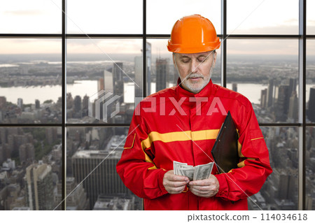 Portrait of construction worker in red uniform is counting money. Urban cityscape background. Portrait of construction worker in red uniform is counting money. Urban cityscape background. 114034618